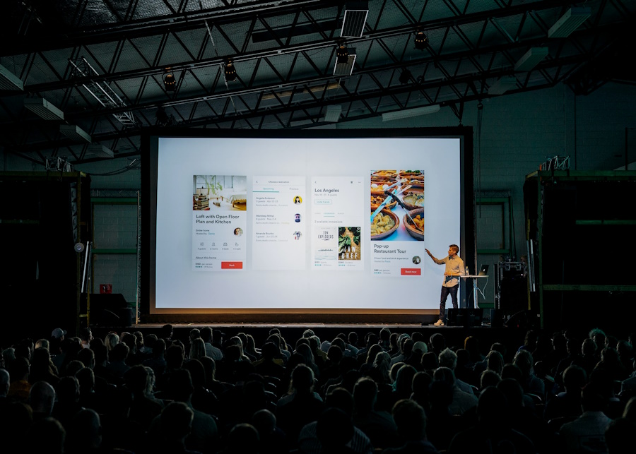 A presentation on a monitor being narrated by a presenter to a large crowd.